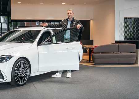 Person standing beside a white Mercedes-Benz sedan in a showroom, holding the driver's door open, with a modern interior in the background.