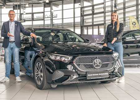 A man and woman stand beside a shiny black Mercedes-Benz in a bright car showroom.