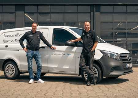 Two men in black shirts stand beside a white Mercedes-Benz van parked in front of a garage with large windows.