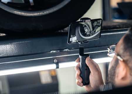 Person holding a smartphone on a stabilizer, recording a close-up of a car tire on a lift in a garage setting.