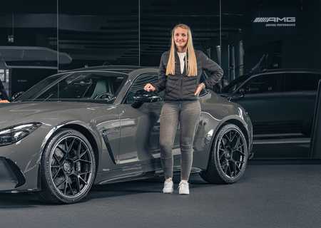 Three people stand beside a sleek gray sports car in a showroom, with AMG branding visible in the background.