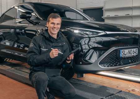 A person in a black jacket kneels beside a black car on a lift in a garage, smiling and holding a tool.
