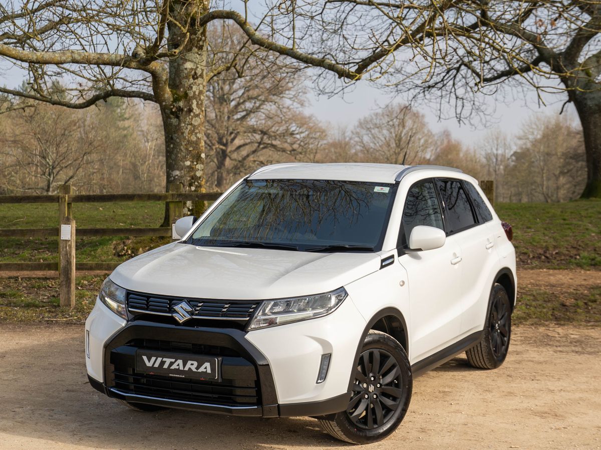 White Suzuki Vitara SUV parked on a dirt path, with leafless trees and a wooden fence in the background under a clear sky.