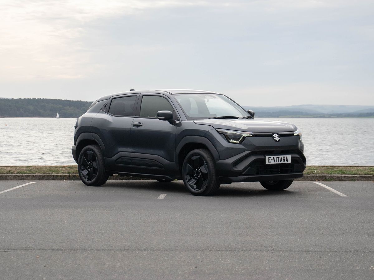 A sleek e VITARA parked by a waterfront under a cloudy sky, with a scenic view of distant hills across the water.