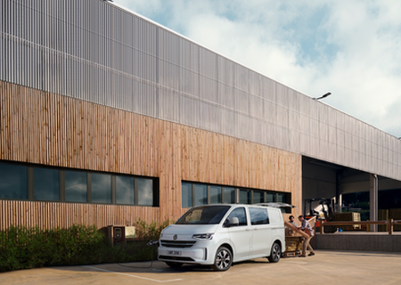 A white van parked in front of a modern building with wooden siding. People are loading or unloading items from the back of the van.