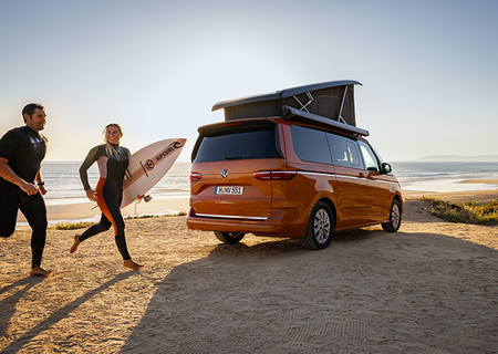 orange camper van on a beach at sunset, one holding a surfboard.