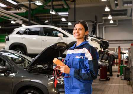 A smiling mechanic in a blue uniform holds diagnostic tools in a garage with several cars, including one with an open hood.