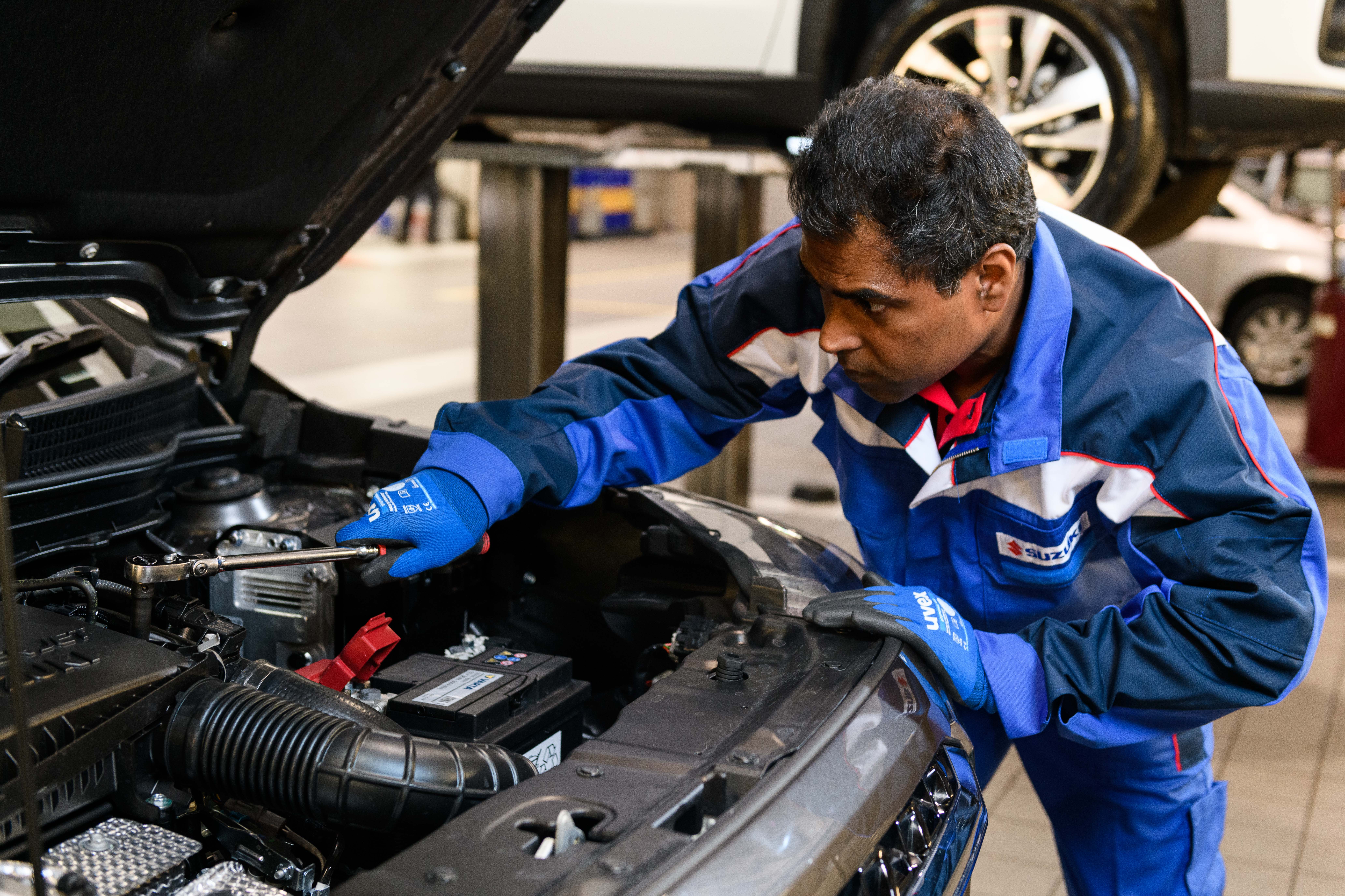 A mechanic in blue uniform inspects a car engine in a garage, using a tool to adjust components.