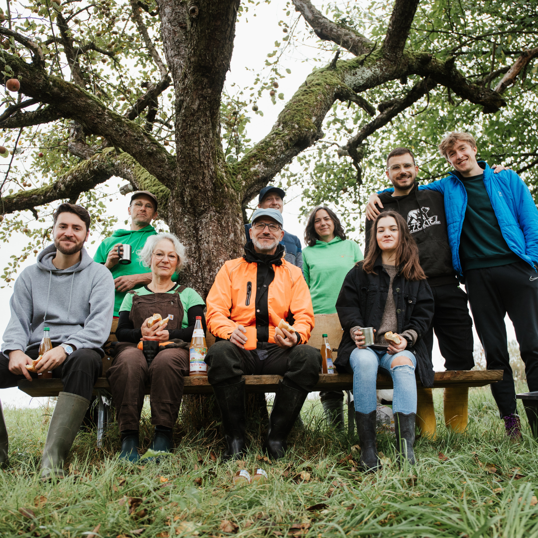 Eine Gruppe von Menschen sitzt auf einer Bank unter einem Baum, genießt Getränke und Snacks und trägt legere Outdoor-Kleidung und Stiefel.