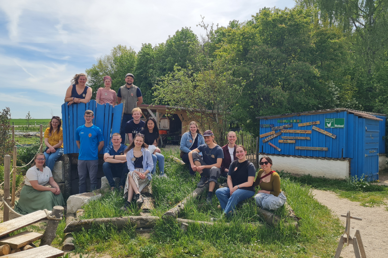 Eine Gruppe von Menschen, die im Freien auf einer Rasenfläche in der Nähe eines blauen Holzgebäudes posiert, im Hintergrund sind Bäume und ein sonniger Himmel zu sehen.