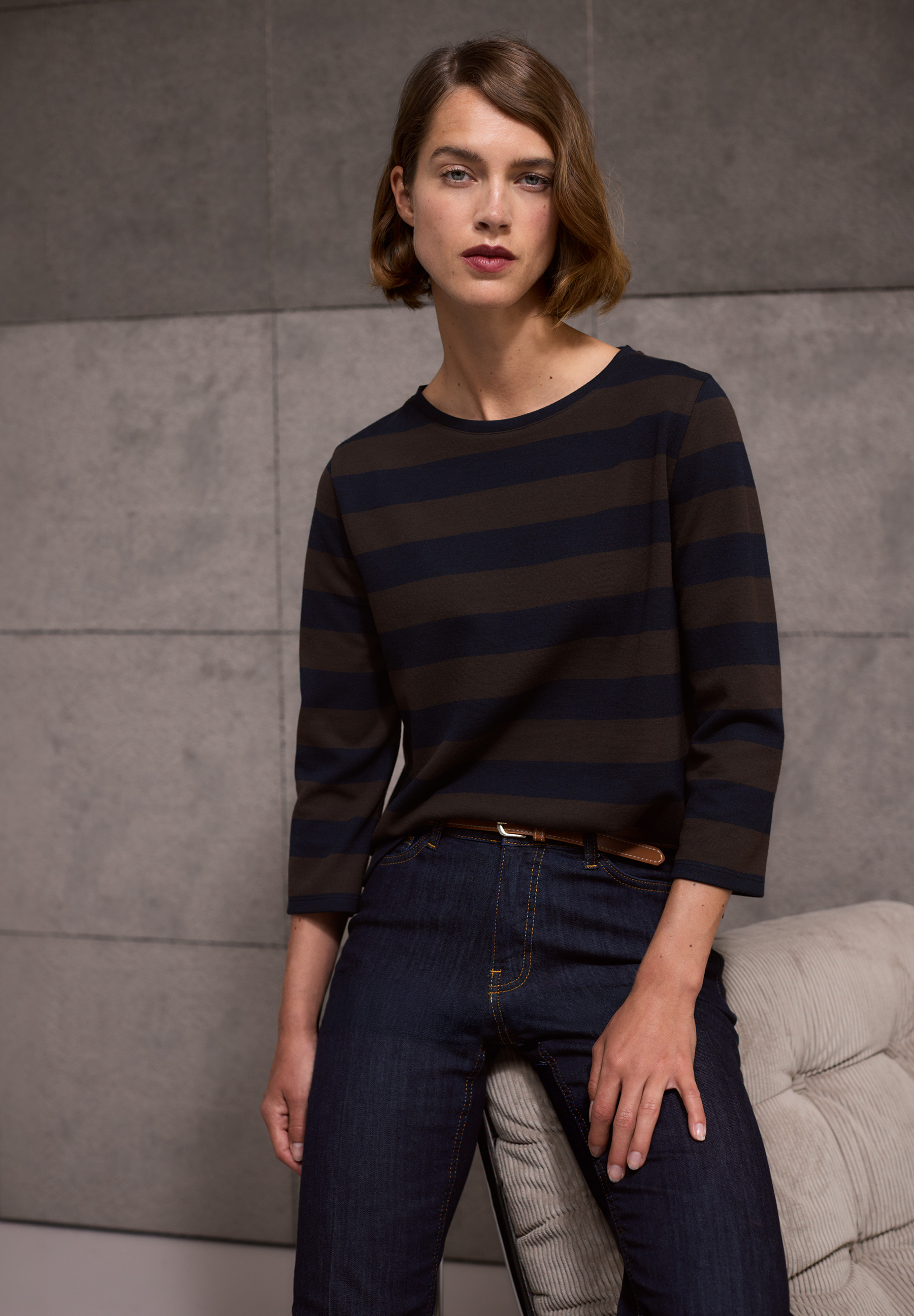 A person with short brown hair wearing a black and brown striped top and dark jeans, leaning on a cushioned chair in a gray room.
