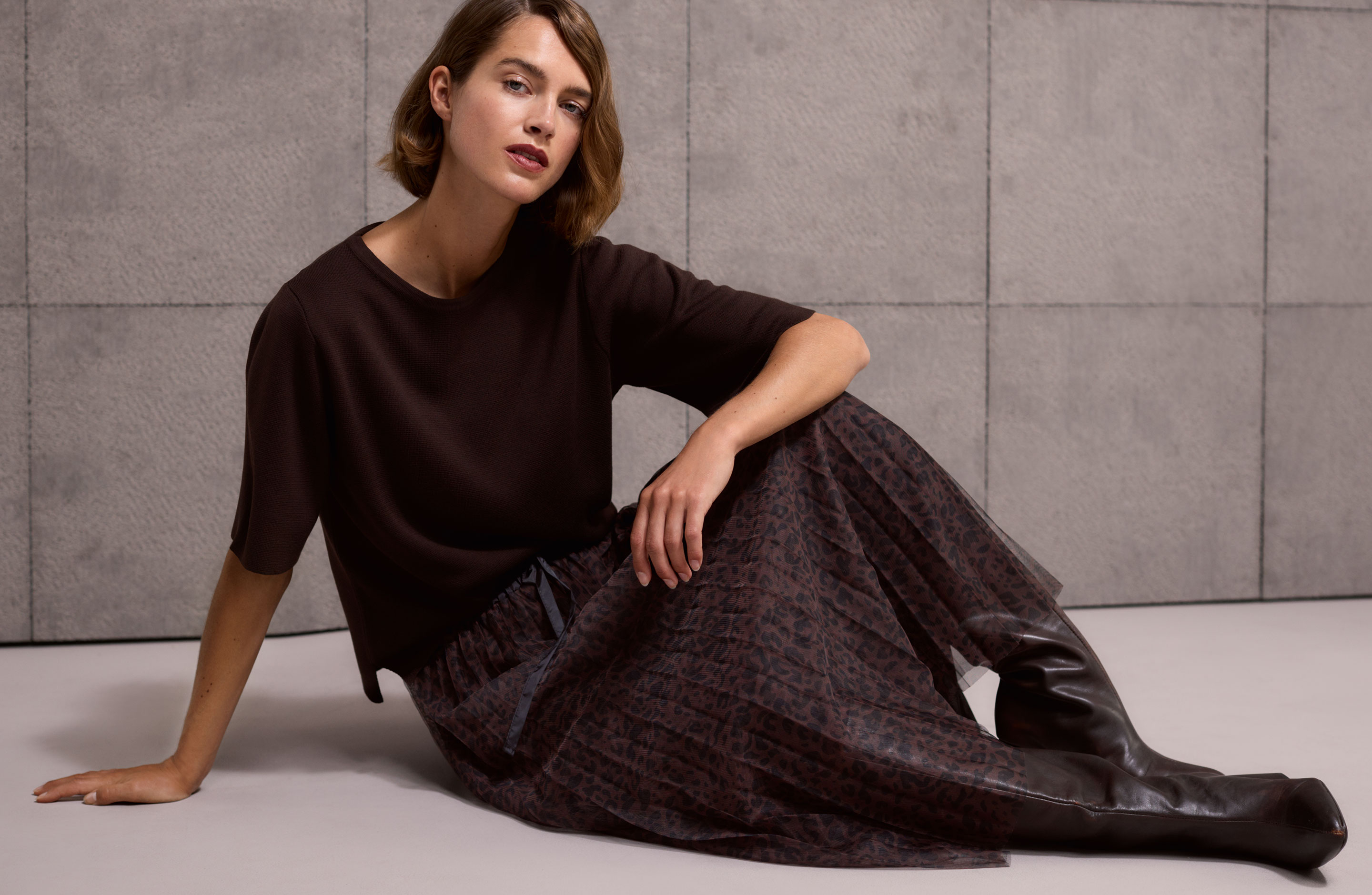 Woman sitting on a floor, wearing a dark top, a patterned skirt, and brown knee-high boots against a textured gray wall.