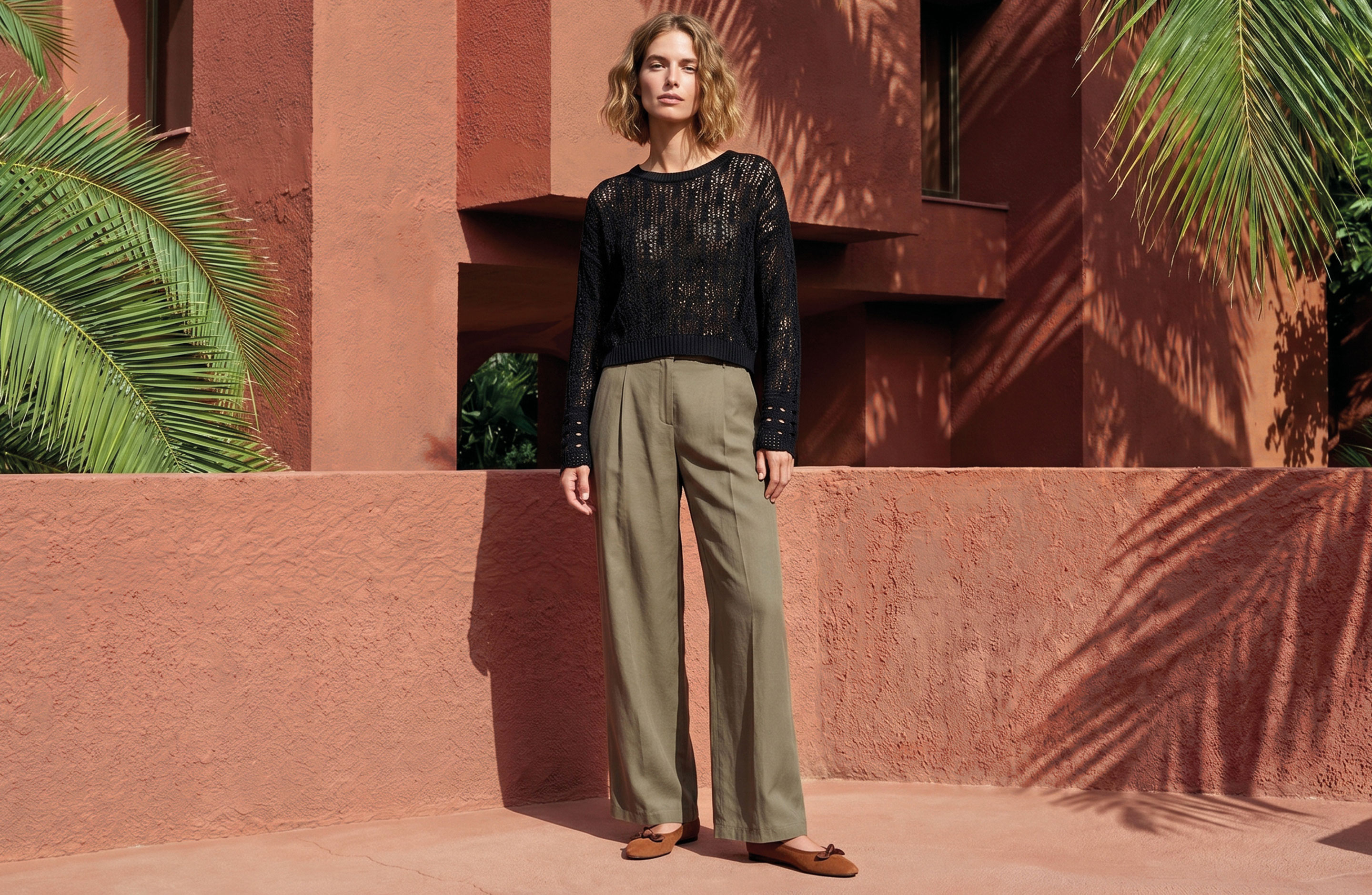 Woman in a black knit sweater and khaki trousers stands outdoors by a terracotta building and palm leaves.