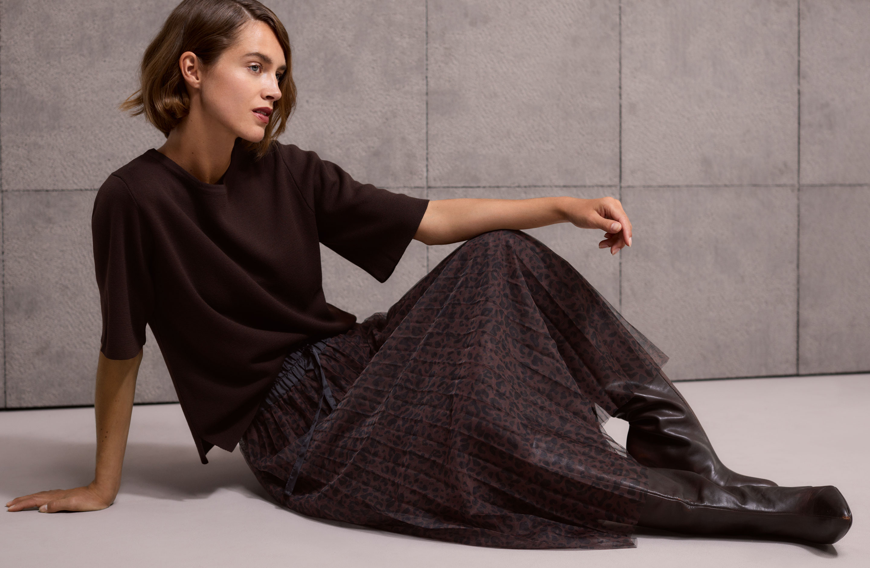 A woman with short hair sits on the floor wearing a dark top, a patterned skirt, and tall brown boots, against a textured gray wall.