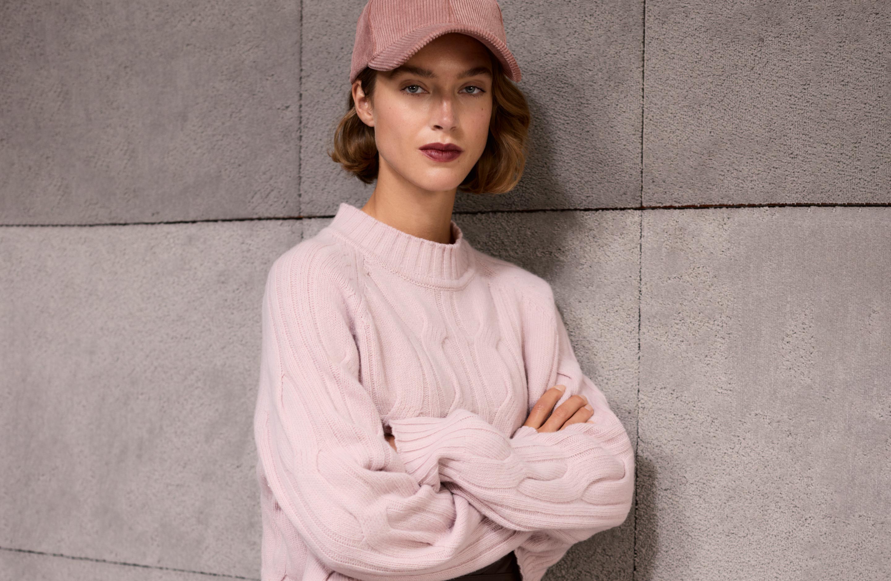 Eine Frau in einem rosa Pullover und einer Mütze, die mit verschränkten Armen vor einer grauen, strukturierten Wand steht.