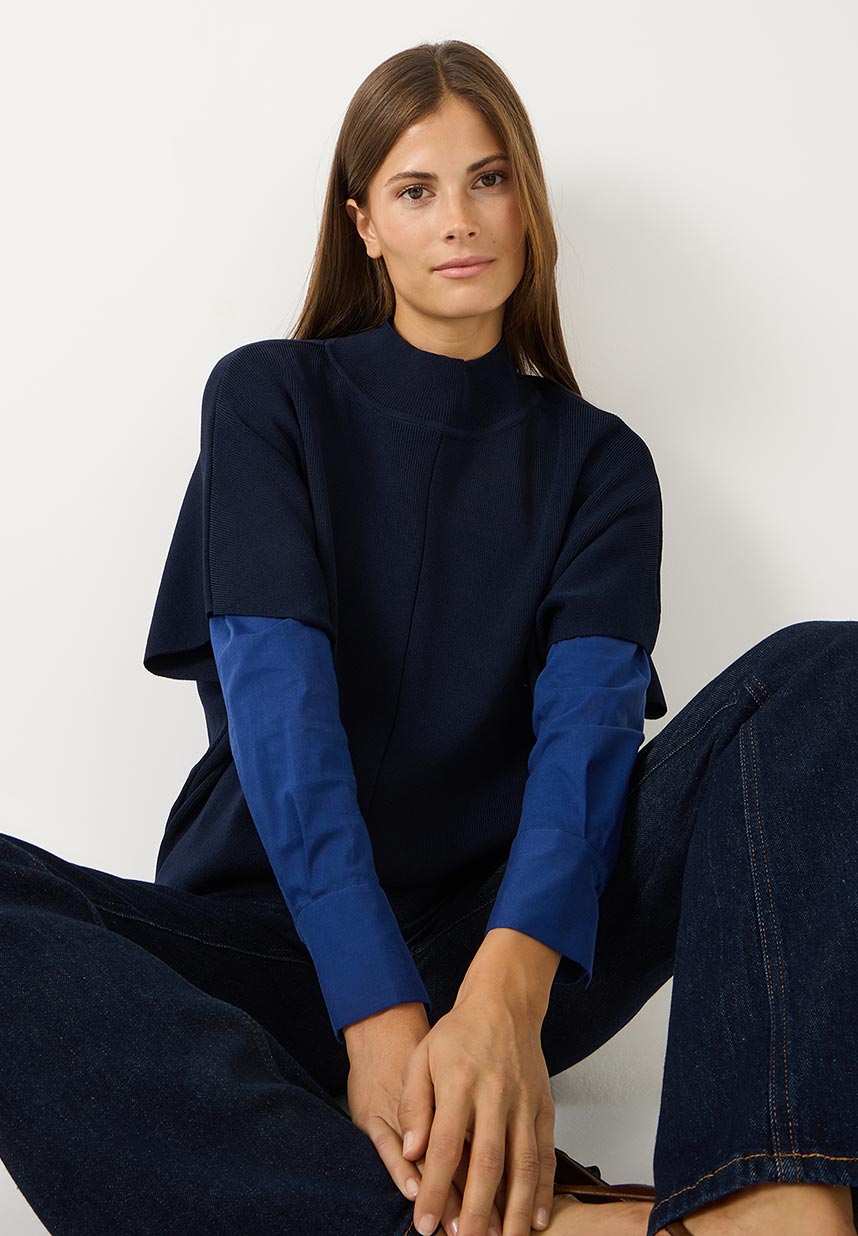 A woman with long brown hair wearing a navy and blue layered top and jeans sits casually on the floor against a white background.