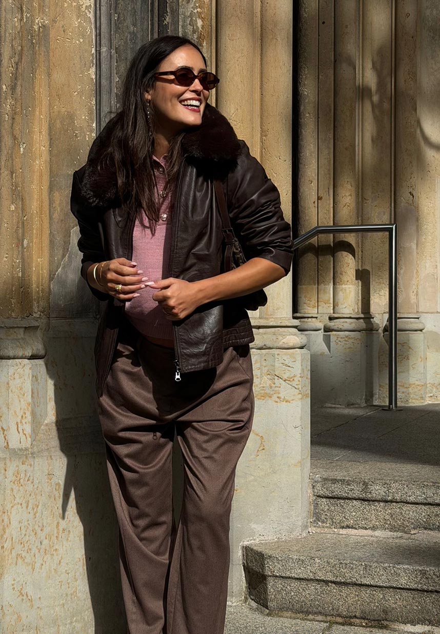 Eine Frau lächelt in der Sonne; sie trägt eine Sonnenbrille, eine braune Lederjacke und eine Hose und steht vor einem Steingebäude.