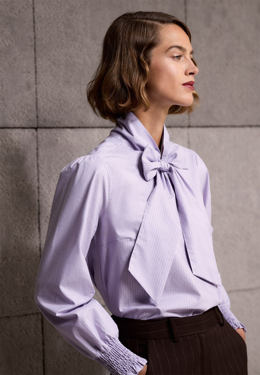 Eine Person mit kurzen Haaren, die eine hellviolette Bluse mit einer großen Schleife am Hals trägt und im Profil vor einer strukturierten Wand posiert.