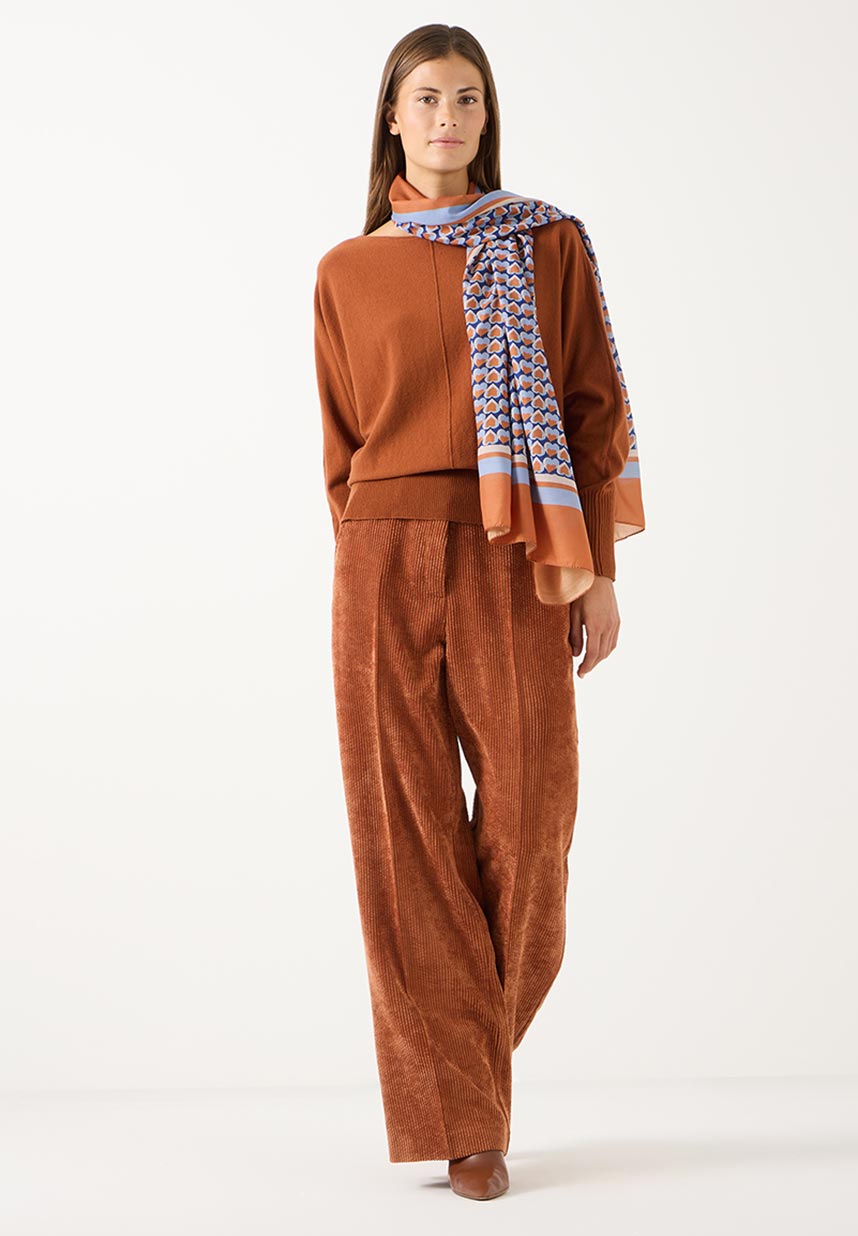 A woman wearing a rust-colored sweater, corduroy pants, and a patterned scarf, standing against a white background.