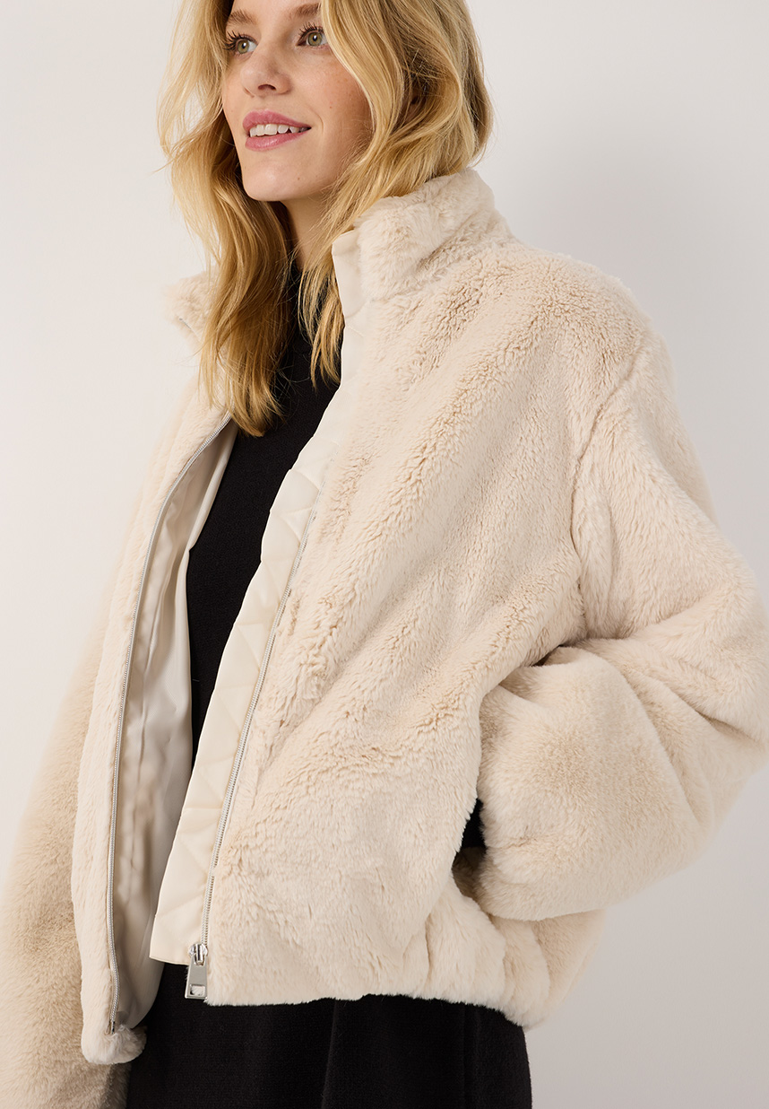 Eine Frau, die einen cremefarbenen Pelzmantel über einem schwarzen Outfit trägt, lächelt, hat langes blondes Haar und steht vor einem schlichten Hintergrund.