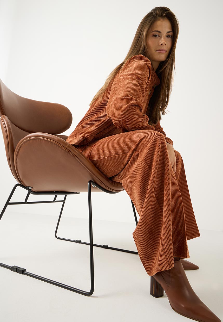 A woman with long brown hair sits in a brown modern chair, wearing a rust-colored corduroy outfit and brown boots, against a white background.