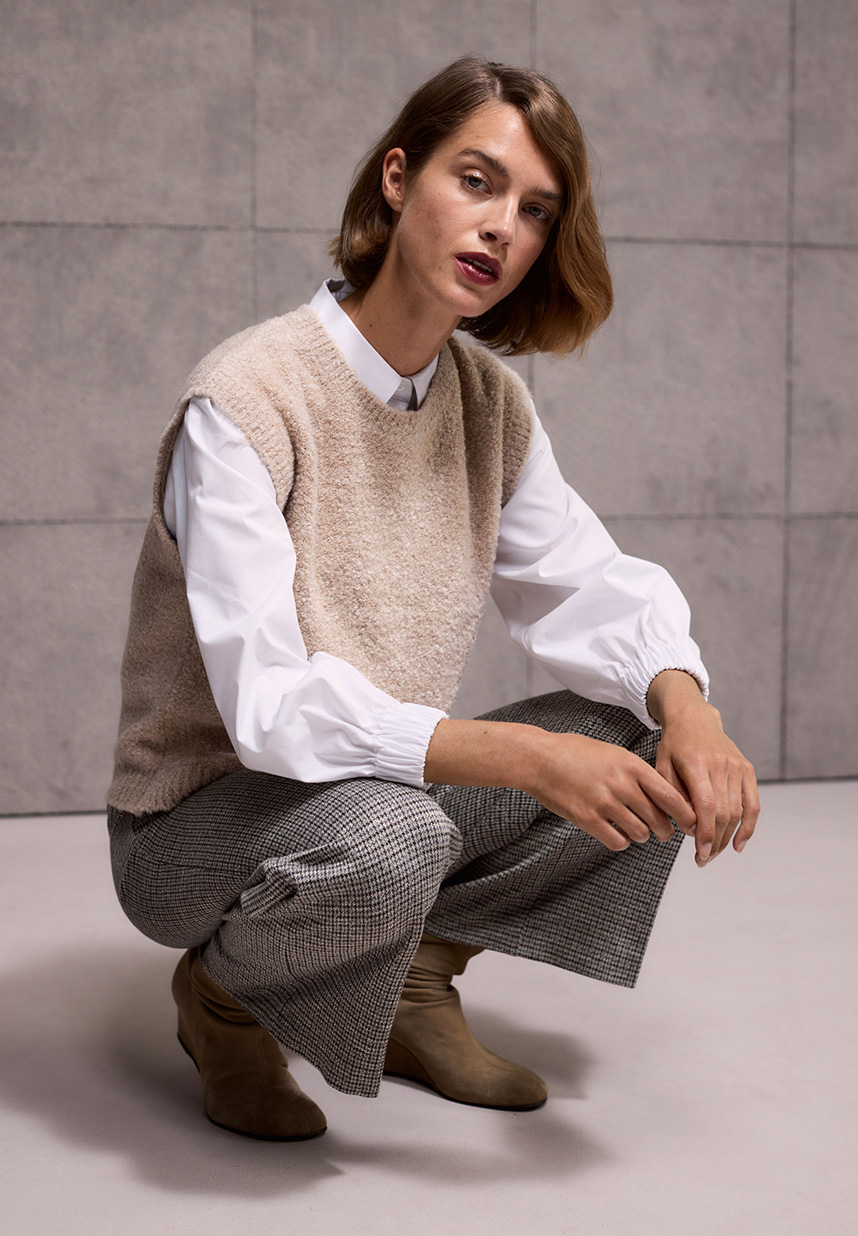 Eine Person mit kurzen braunen Haaren, die eine beige Strickweste über einem weißen Hemd und einer grau gemusterten Hose trägt und vor einem gefliesten Hintergrund steht.