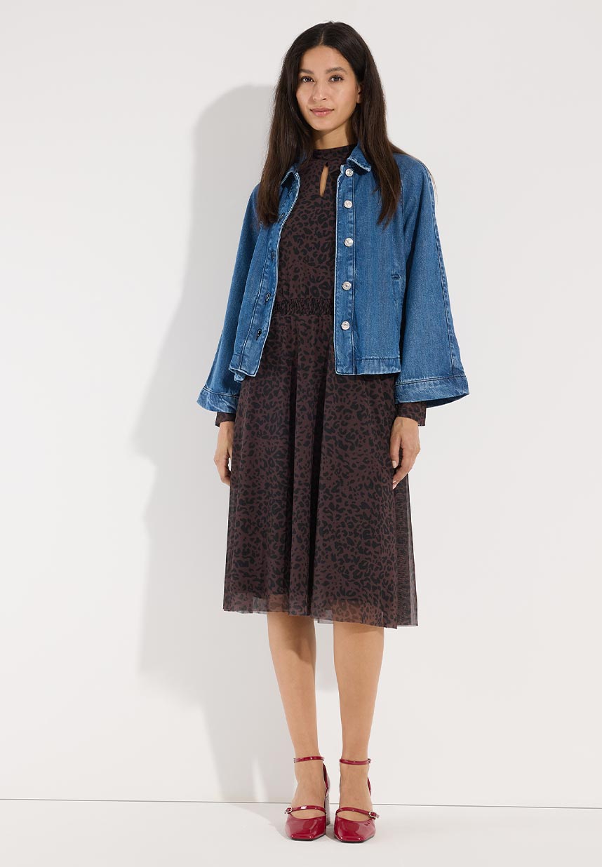 Woman in a dark leopard print dress and blue denim jacket, wearing red heels, standing against a plain background.