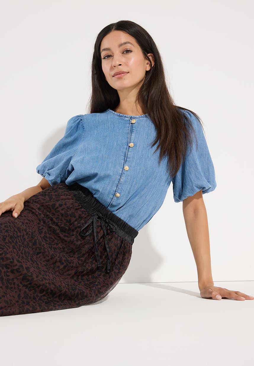 A woman with long dark hair wearing a blue buttoned blouse and patterned skirt leans against a white surface, looking at the camera.