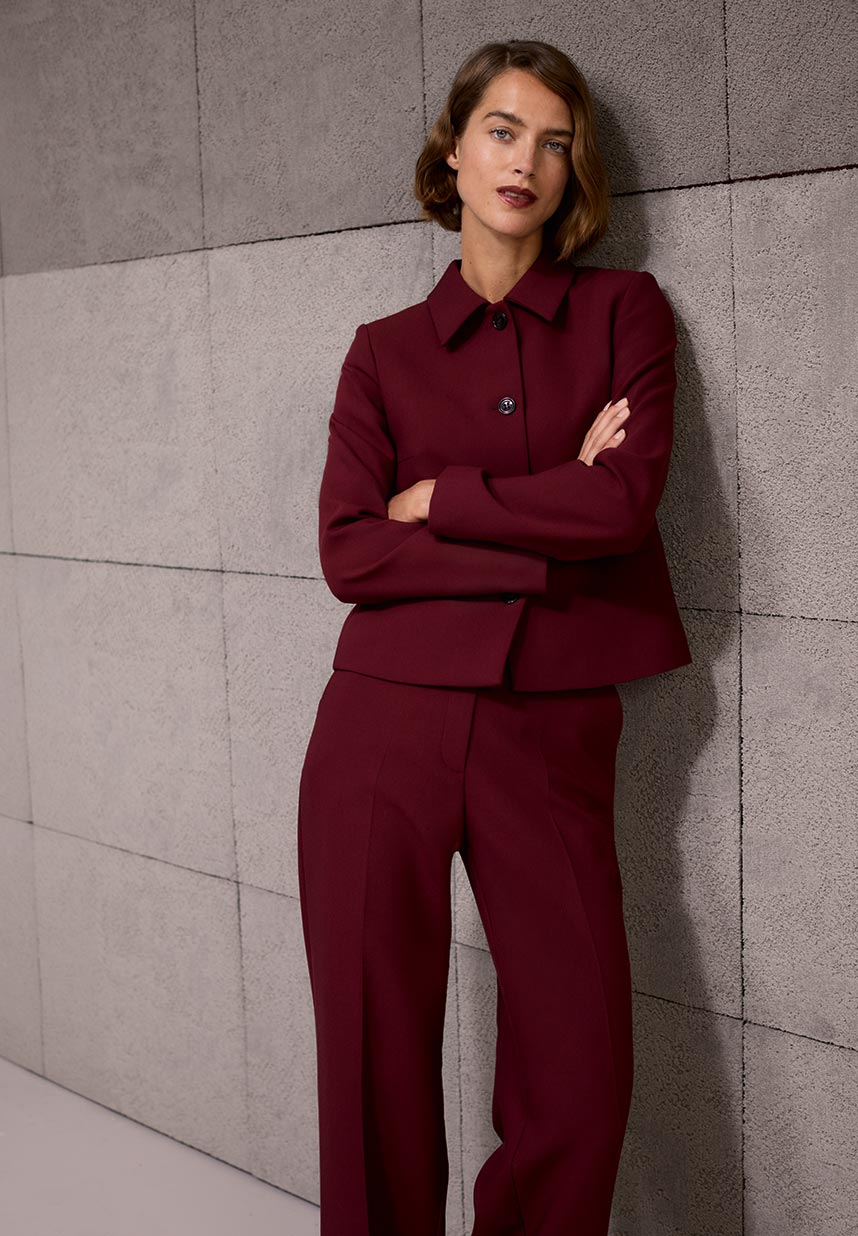 Woman with short brown hair wearing a burgundy suit stands against a textured gray wall, arms crossed, with a confident expression.
