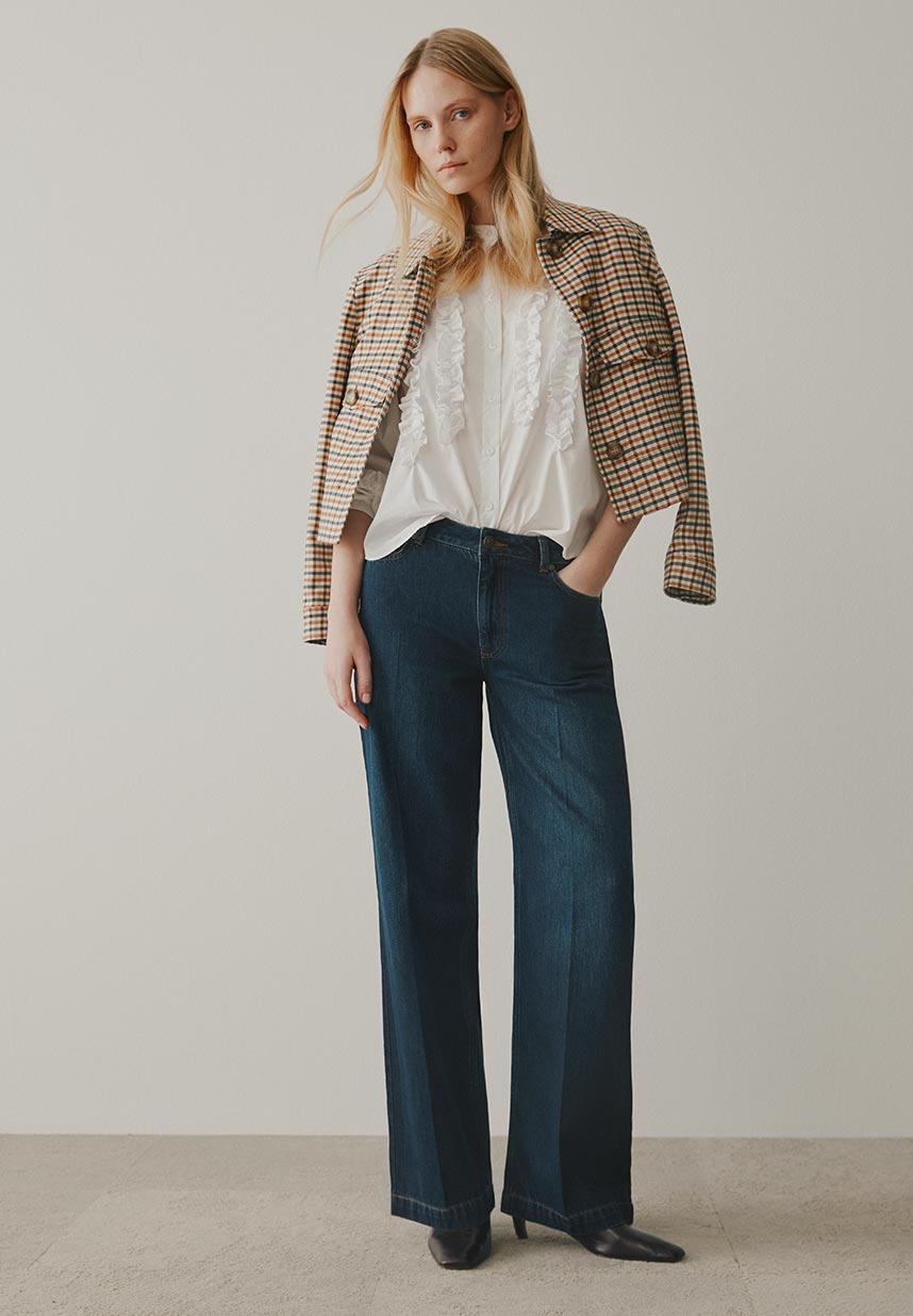 A woman stands against a plain background, wearing a plaid blazer, white blouse, and wide-leg jeans, with her hand in her pocket.