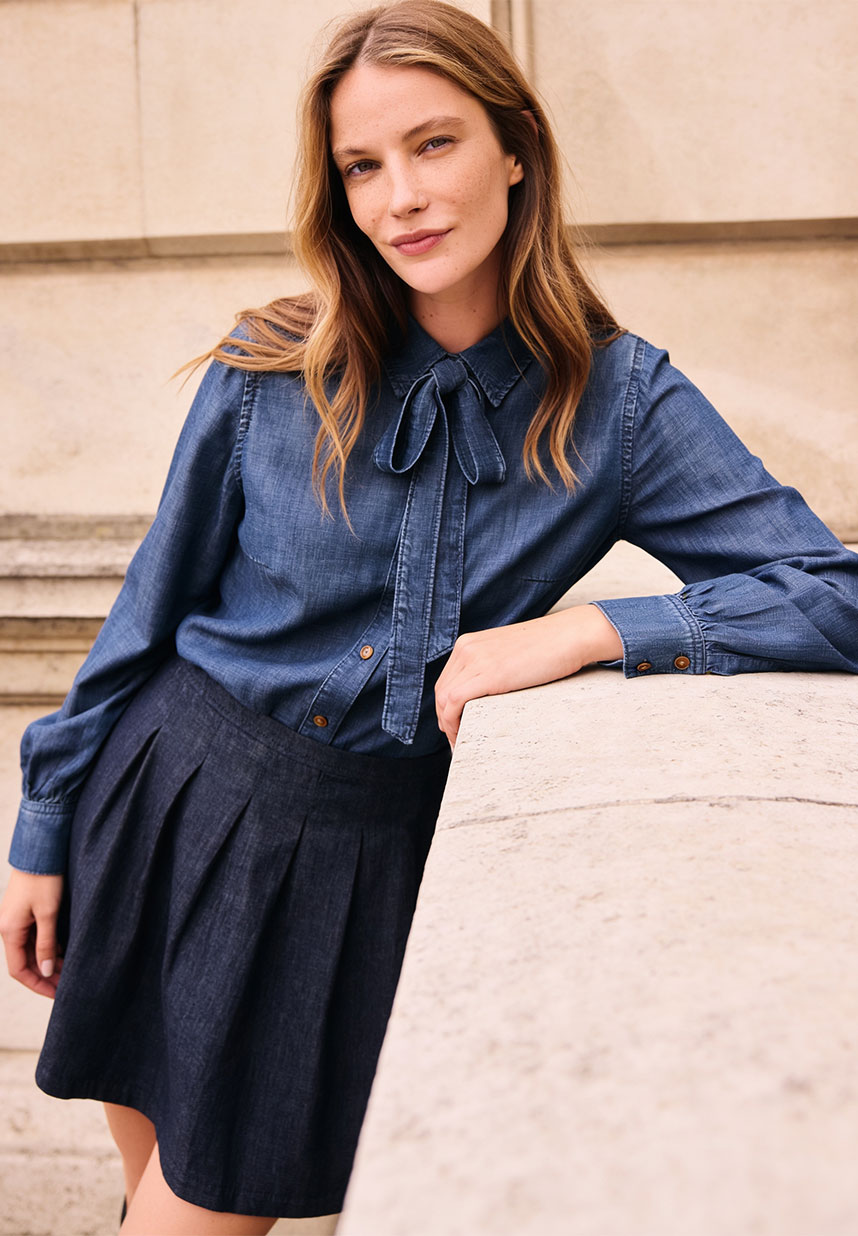 Eine Frau, die eine blaue Jeansbluse mit Schleife und einen Faltenrock trägt, lehnt an einem Steinbalustrade und lächelt sanft.