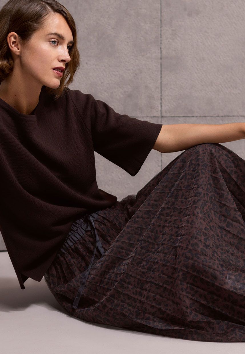 A person with short hair sits wearing a dark top and a patterned skirt, against a gray concrete background.