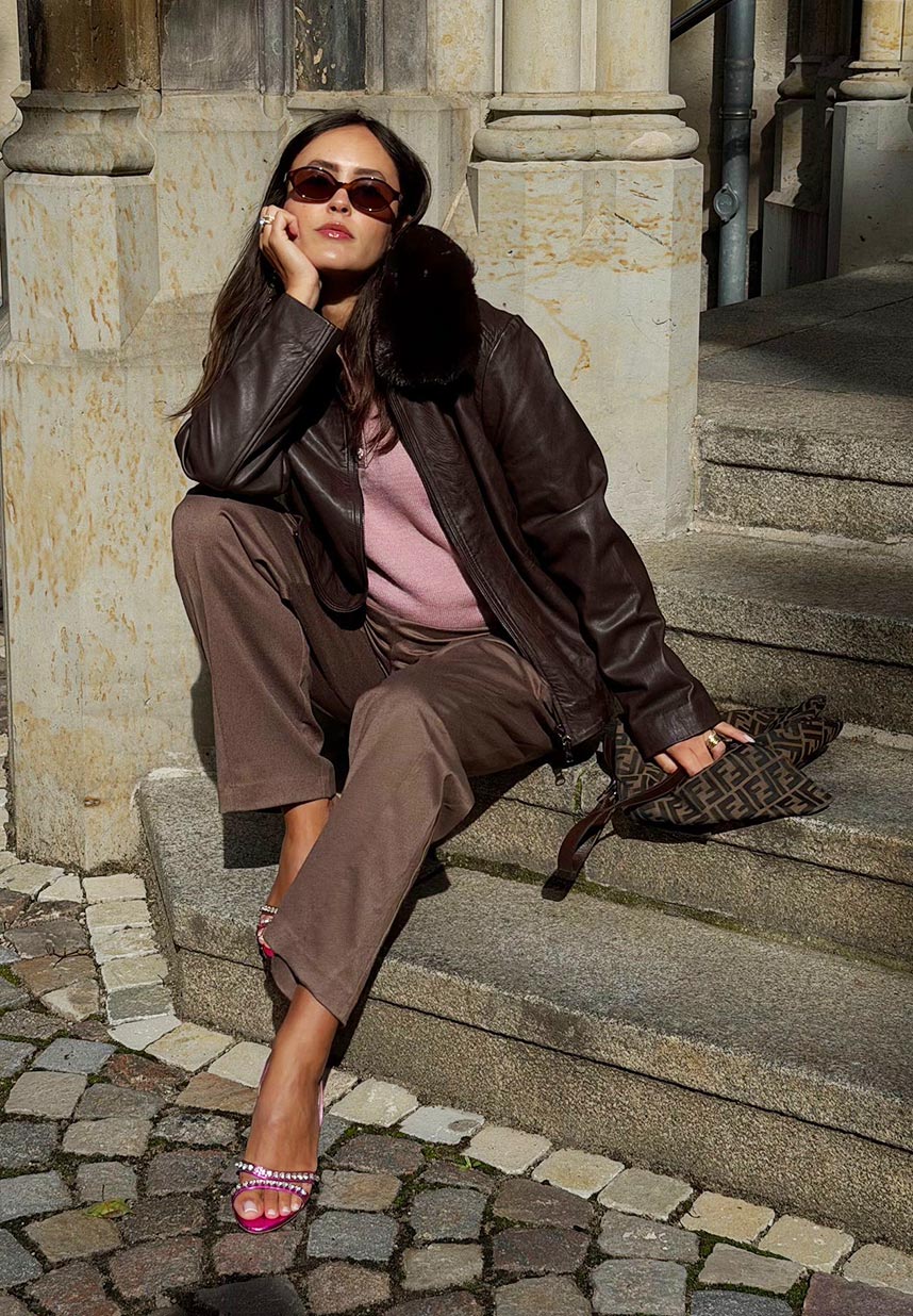 Eine Frau lächelt in der Sonne; sie trägt eine Sonnenbrille, eine braune Lederjacke und eine Hose und steht vor einem Steingebäude.