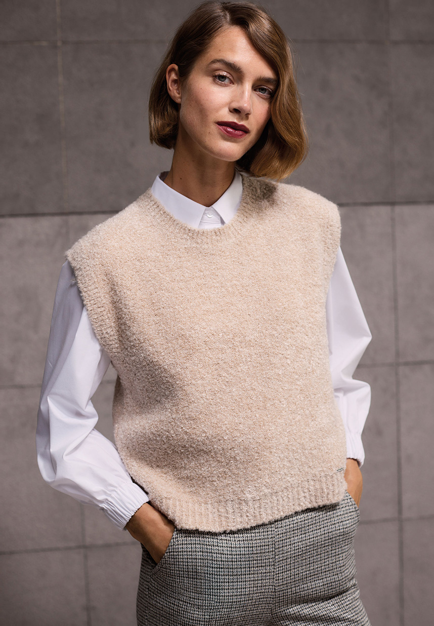 Eine Person mit kurzen braunen Haaren, die eine beige Strickweste über einem weißen Hemd und einer grau gemusterten Hose trägt und vor einem gefliesten Hintergrund steht.