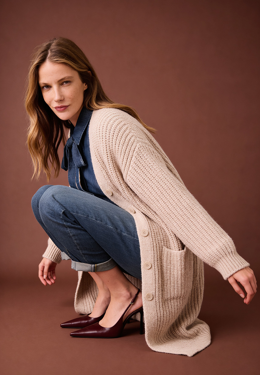 Frau hockt und trägt eine Jeans und eine Denim Bluse mit einem langen Strickmantel