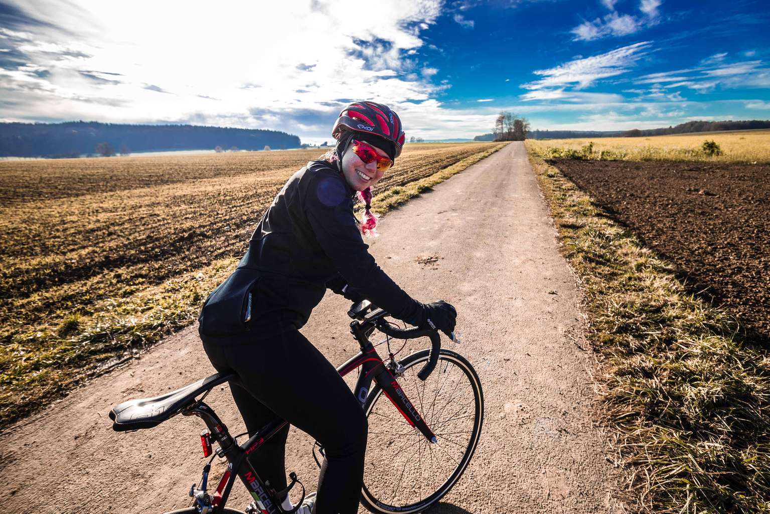 Rennradfahrerin blickt in die Kamera. Darum herum Felder und blauer Himmel.
