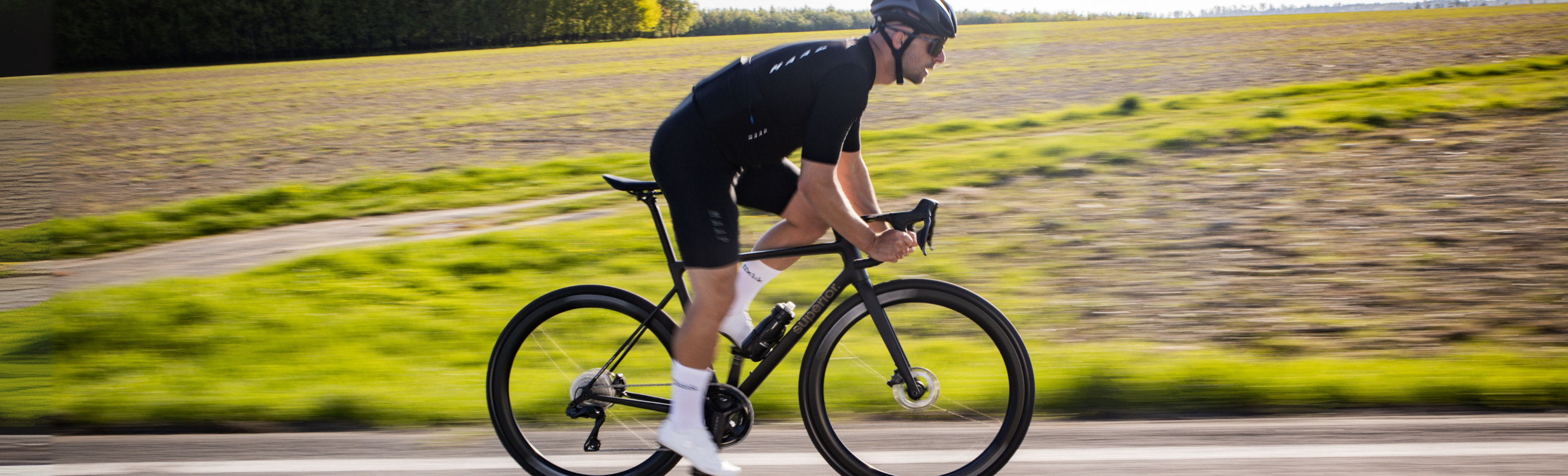 Ein Radfahrer mit schwarzer Fahrradbekleidung und Helm fährt an einem sonnigen Tag mit einem Rennrad auf einer gut geteerten Landstraße. Aufgrund der Geschwindigkeit und der Bewegung, in der das Bild aufgenommen wurde, ist der Hintergrund verschwommen. Dieser zeigt grüne und braune Felder.