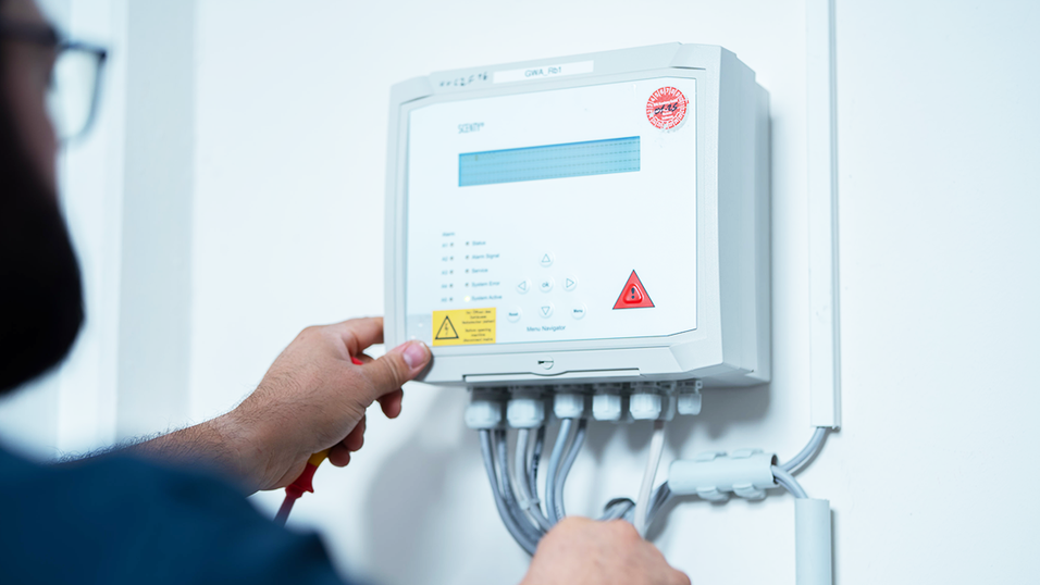 A person adjusting or inspecting a white security or gas detection system mounted on a wall, with several cables connected at the bottom.