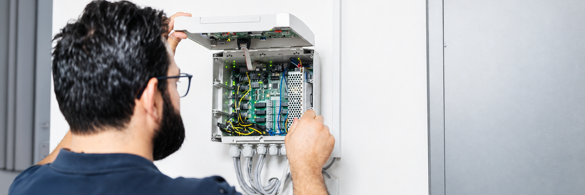 A man in glasses examines an open enclosure with electronics with circuit boards and wiring, mounted on a white wall.