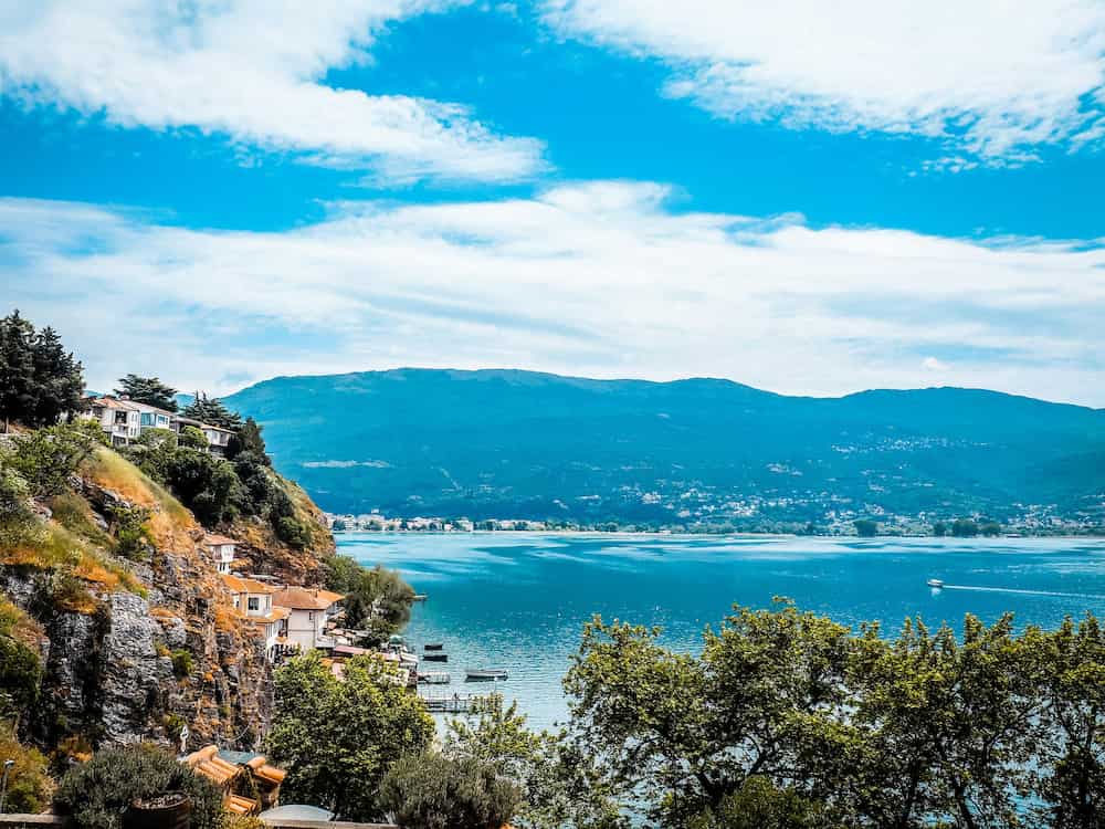 Cliffside houses overlooking a pristine blue lake with distant mountains and a partly cloudy sky.