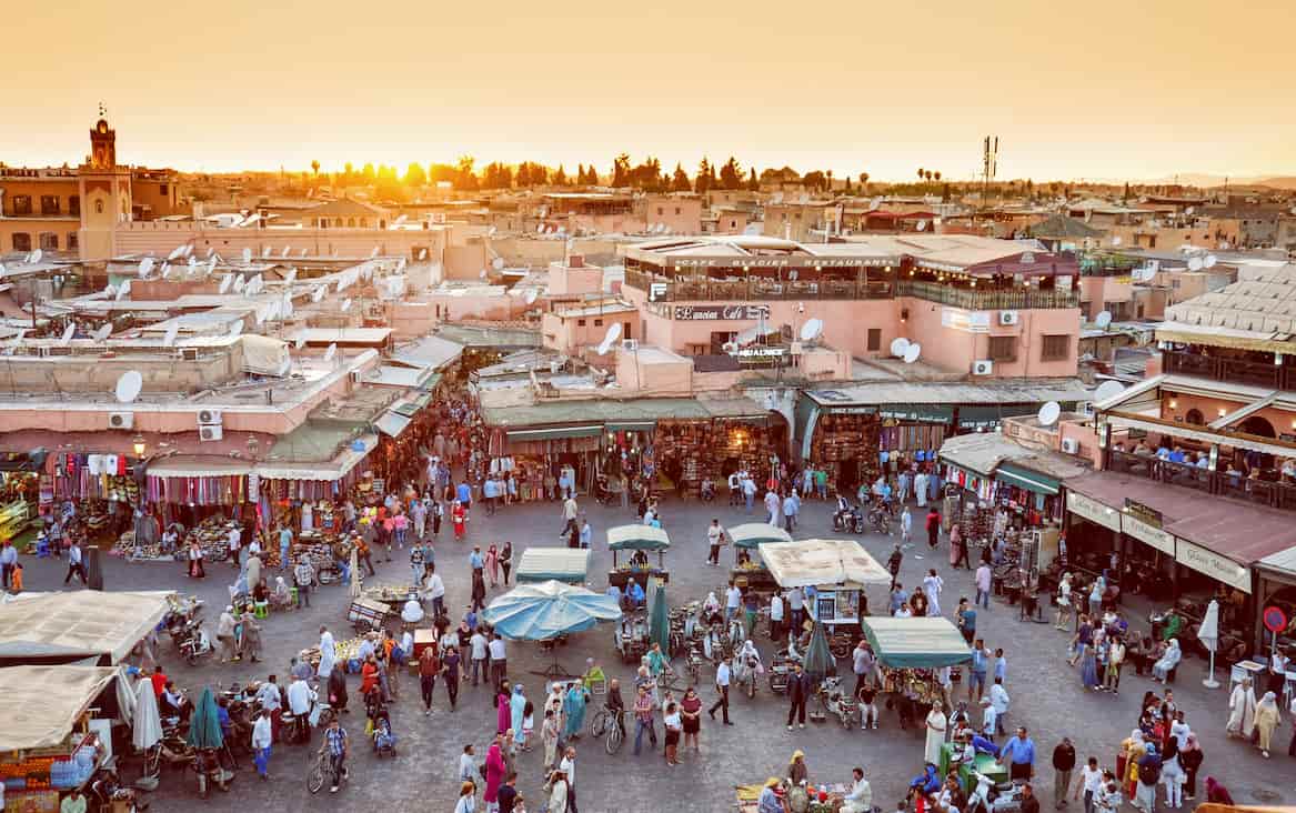 A bustling market scene in Marrakesh at sunset, with vibrant stalls and numerous people navigating the busy square.