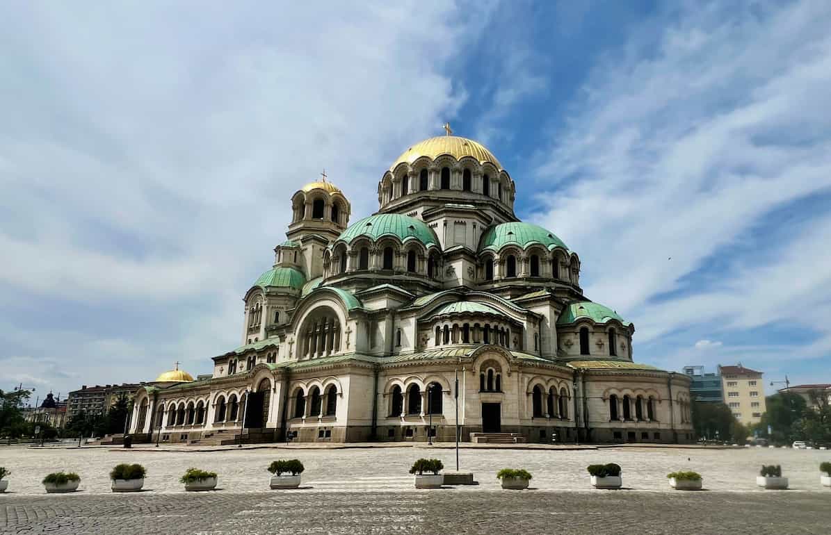 Ornate cathedral with green and gold domes under a blue sky, set in an empty plaza.