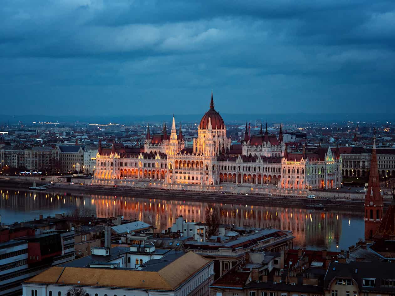 Illuminated Hungarian Parliament building at dusk, reflecting on the Danube River, under a cloudy sky.
