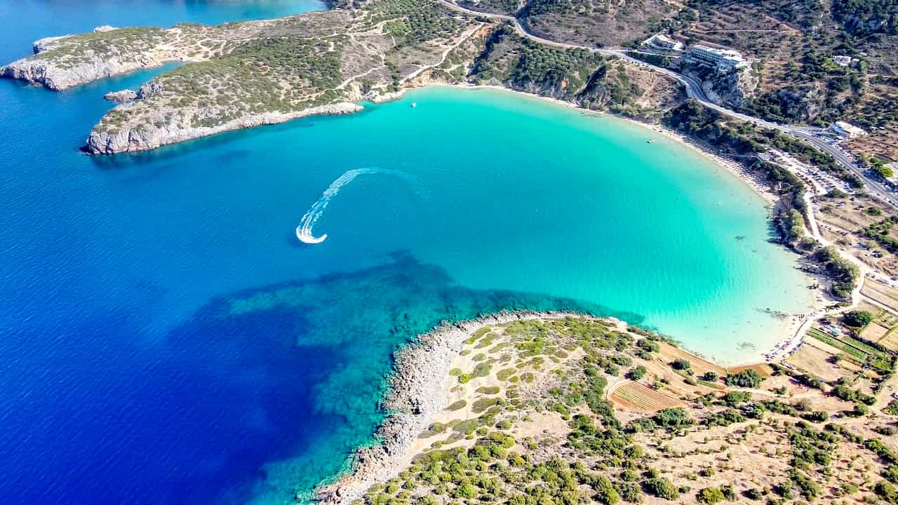 Aerial view of a serene turquoise bay with a speedboat making a curve, surrounded by rugged cliffs and greenery.