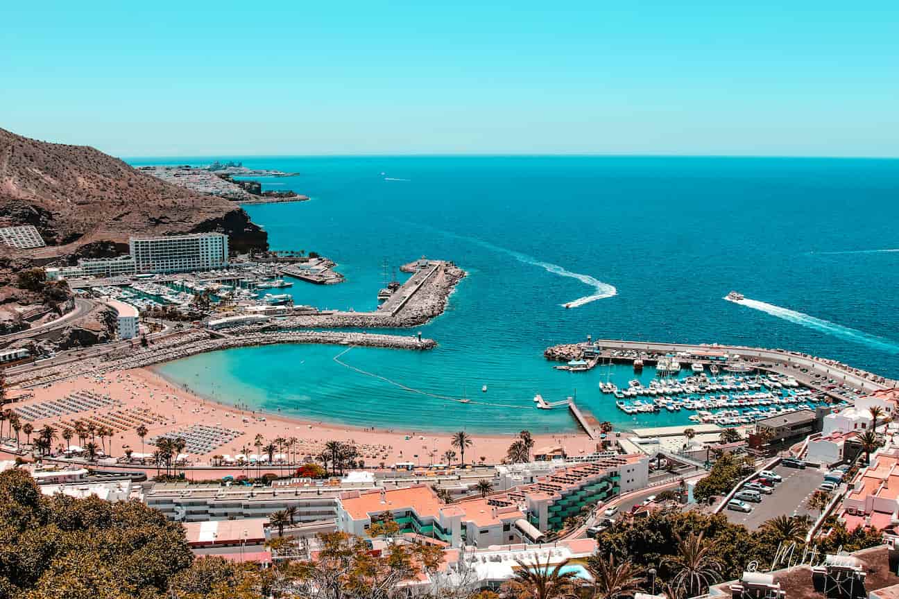 Overlooking a vibrant coastal resort with a curving beach, marina, and hotels, surrounded by rugged hills and a clear blue sea under a sunny sky.