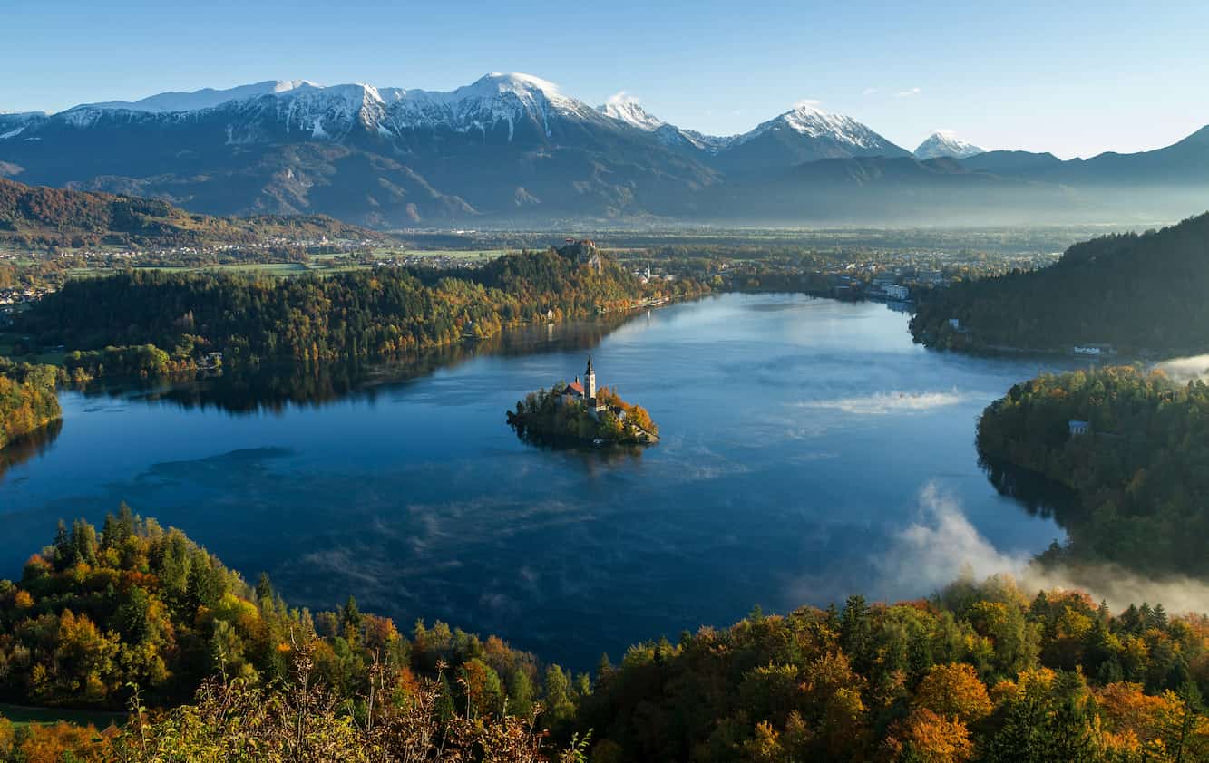 A serene lake surrounded by colorful autumn forests, an island with a church in the center, and snow-capped mountains in the background under a clear sky.