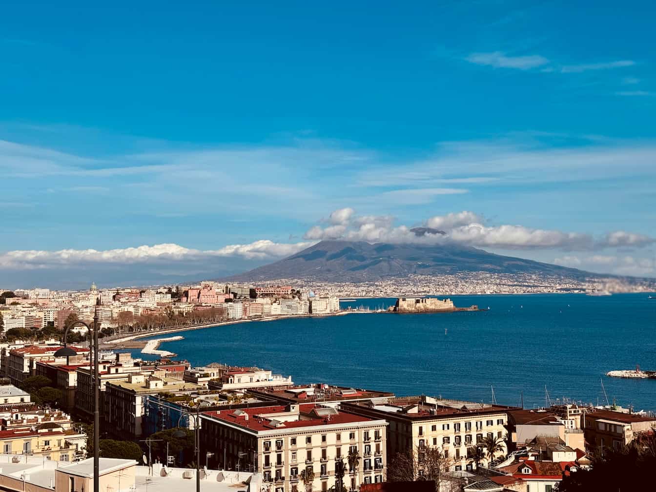 Coastal cityscape with buildings lining the shore, expansive blue sea, and a mountain in the background under a clear blue sky.