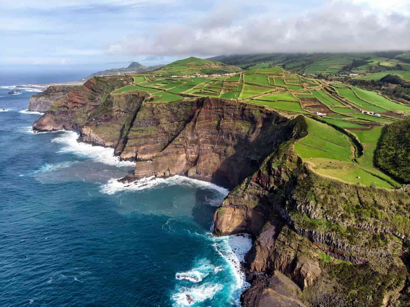 Clifftop fields overlooking a rugged coastline with ocean waves crashing against the rocky shore, under a partly cloudy sky.