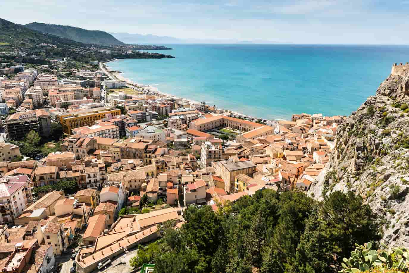 Aerial view of a coastal town with terracotta-roofed buildings by a vibrant blue sea and lush green hills in the background.