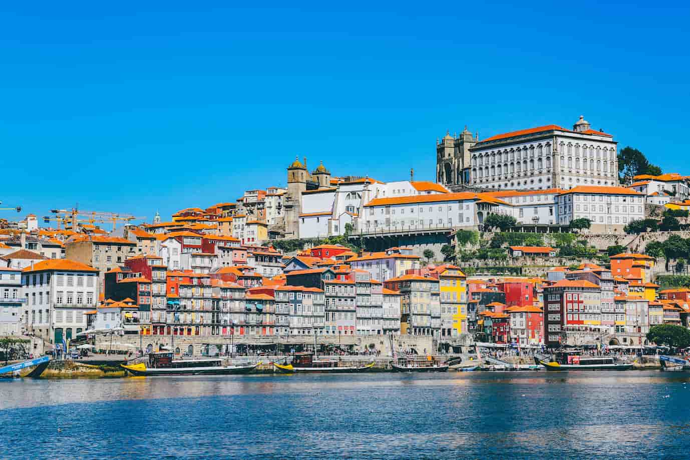 Colorful hillside cityscape with old buildings and orange roofs under a clear blue sky, reflected in a calm river below.